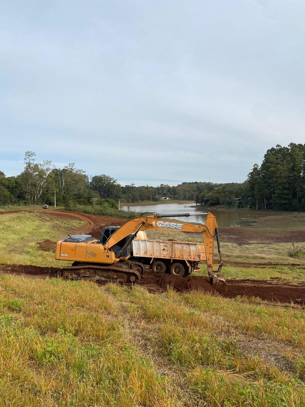 Escavadeira carregando caminhão caçamba durante limpeza de terreno às margens de açude